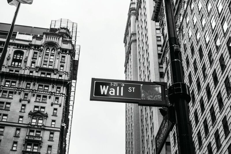 A black and white photograph looking up at a Wall St street sign mounted on a lamppost between tall stone office buildings in lower Manhattan.