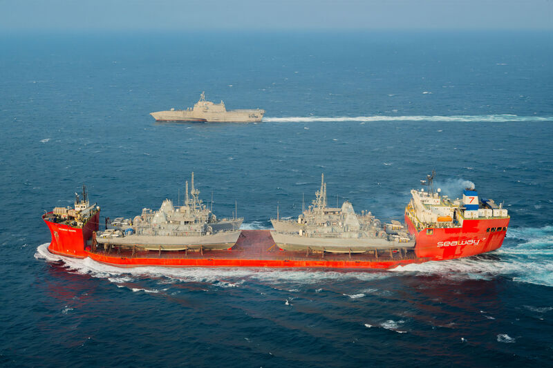 An aerial view of a red heavy-lift merchant ship under way on open blue water, a grey warship in the distance behind it.