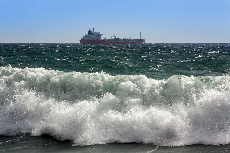 A red oil tanker on the horizon, seen beyond a large breaking wave in the foreground.