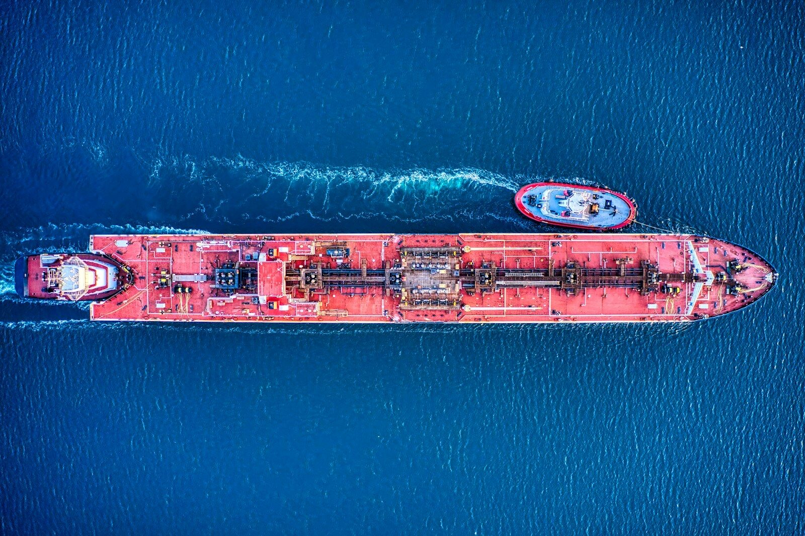 A red oil tanker seen from directly above on dark blue water, with a tugboat alongside.