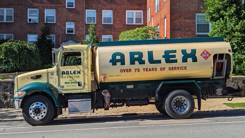 A cream and green heating oil delivery tanker truck parked on a residential street in front of brick apartment buildings.
