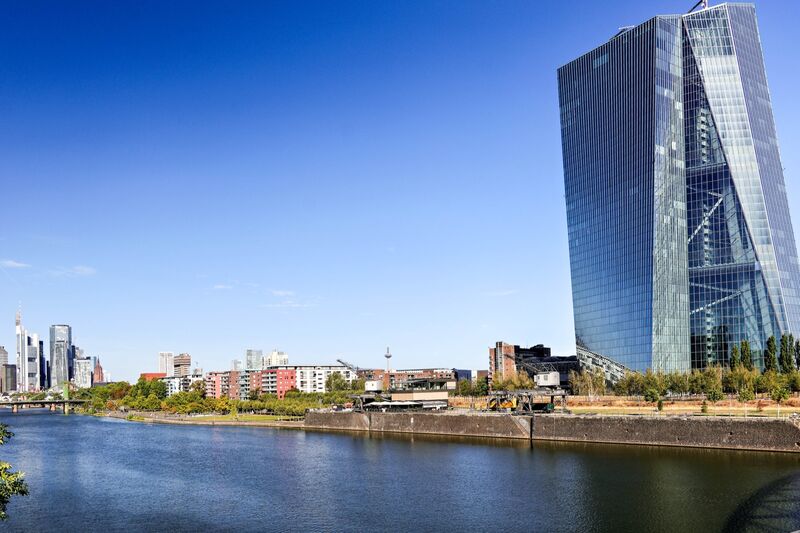 The twin glass towers of the European Central Bank rise beside the River Main in Frankfurt, with the city's skyline in the distance under a clear blue sky.
