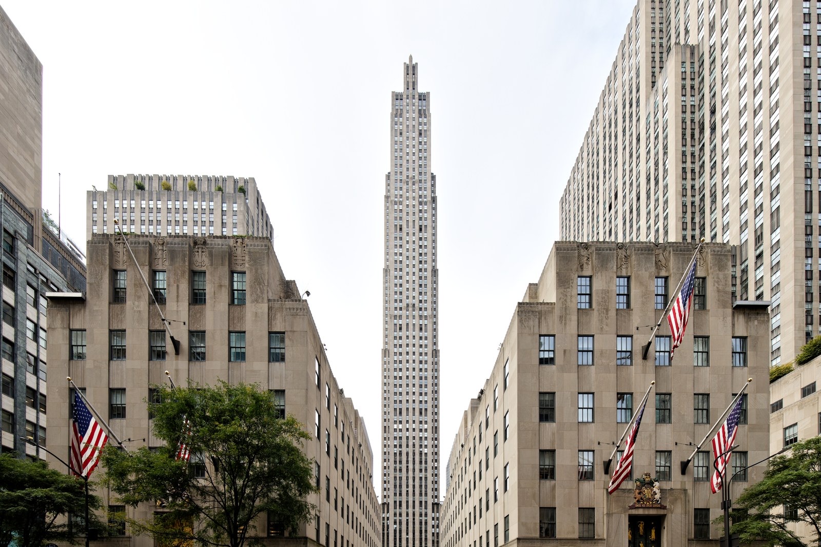 The slender limestone tower of 30 Rockefeller Plaza rising between two lower stone buildings hung with American flags, seen from Fifth Avenue under a pale sky.