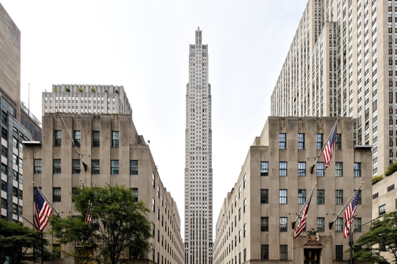 The slender limestone tower of 30 Rockefeller Plaza rising between two lower stone buildings hung with American flags, seen from Fifth Avenue under a pale sky.