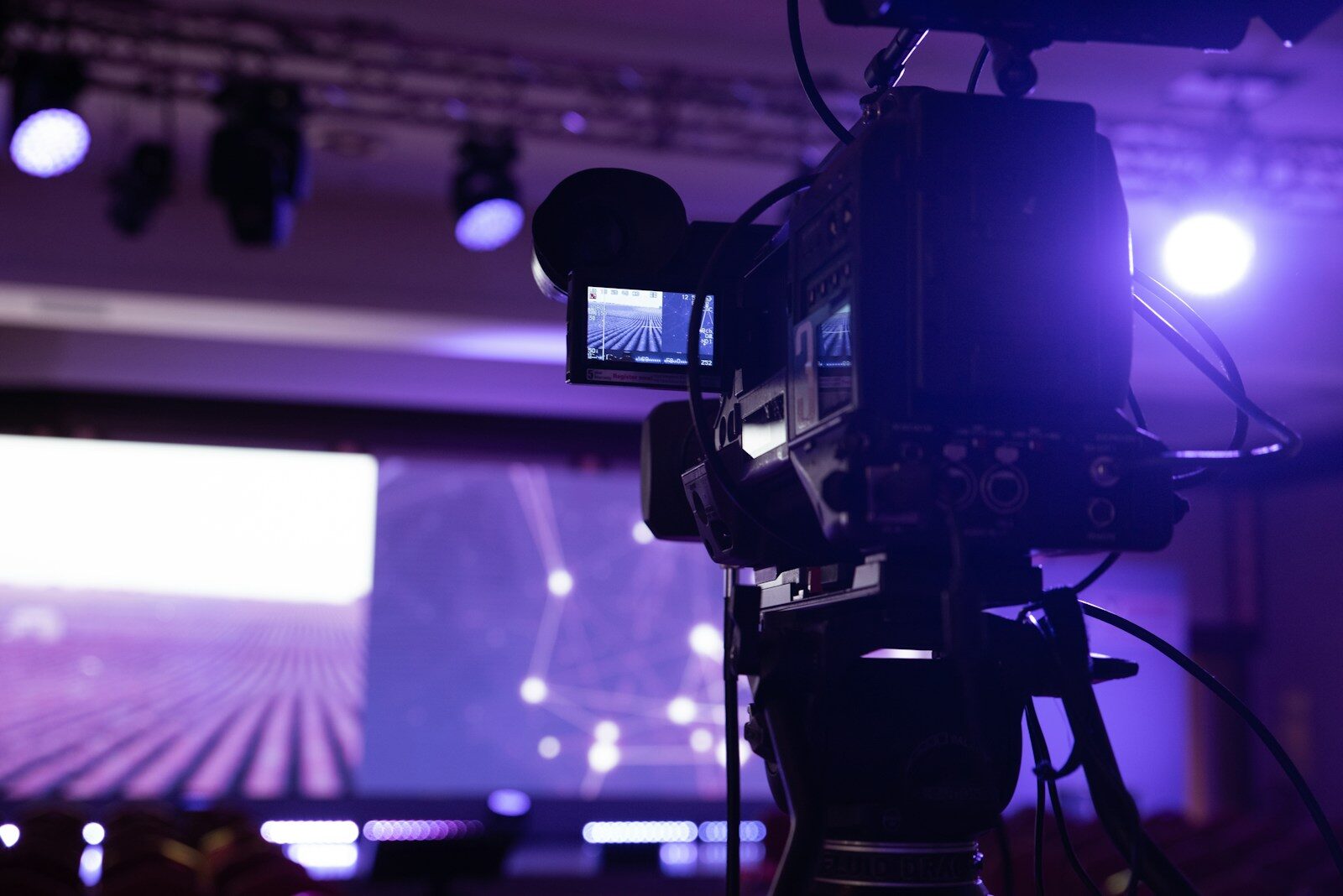 A broadcast video camera on a tripod trained on a lit stage