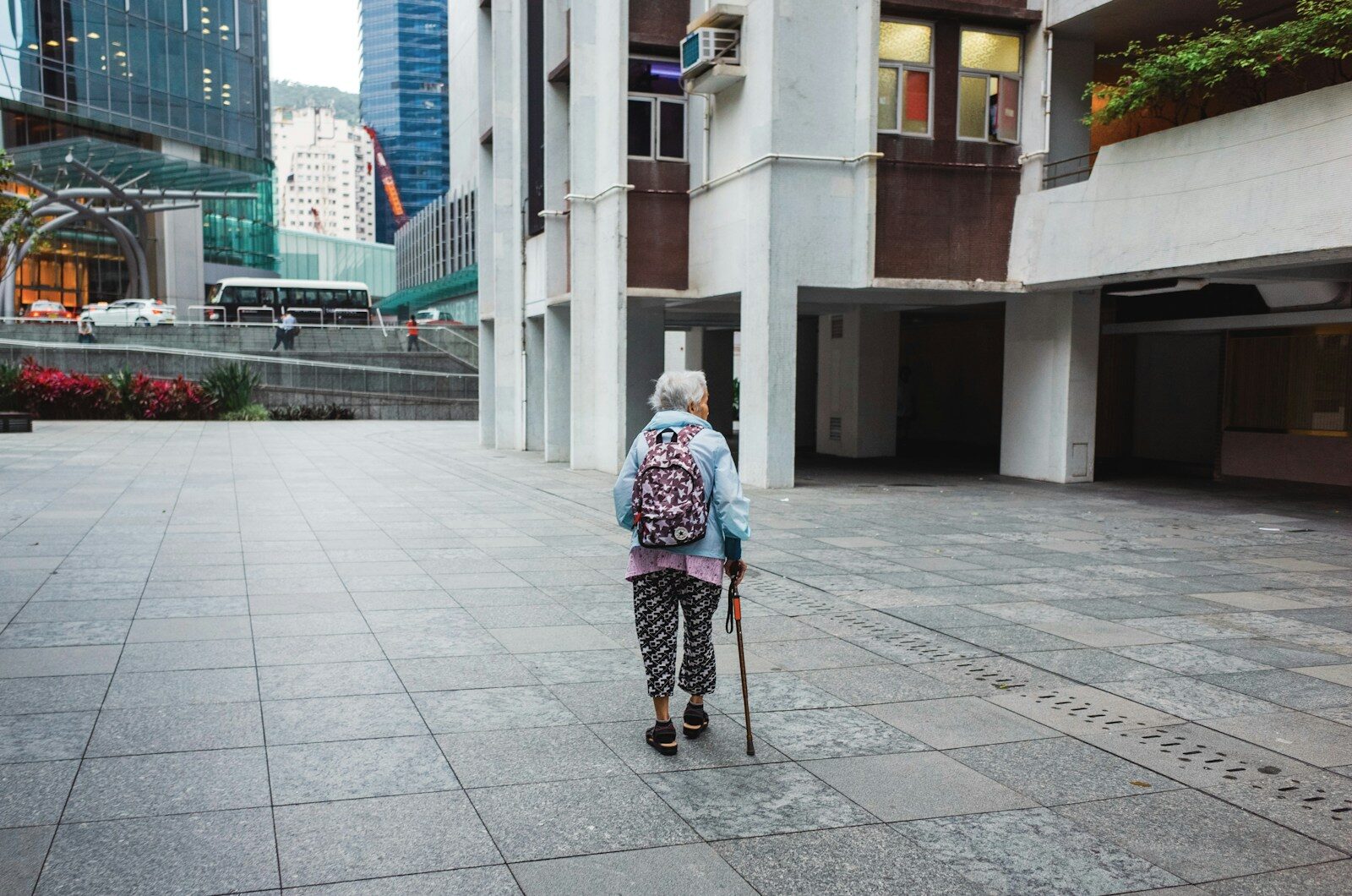 An elderly woman in bright colors and patterned clothes walks across a square in Hong Kong, beside an apartment block. She holds a walking stick. A busy urban environment and traffic is visible in the distance.
