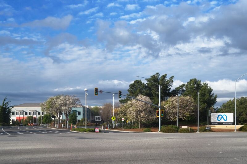The entrance to Meta's headquarters campus at 1 Hacker Way in Menlo Park, California, with the Meta logo sign beside the road, office buildings and flowering trees under a cloudy blue sky.