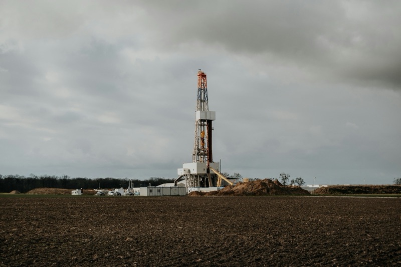 A tall land drilling rig with equipment and soil piles at its base stands in a bare ploughed field under heavy grey clouds.