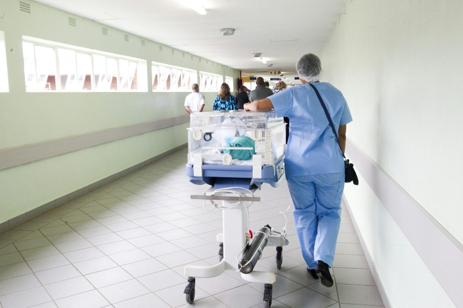 A health worker in blue scrubs and a cap pushes a hospital incubator trolley down a long, bright corridor, with other people walking ahead.