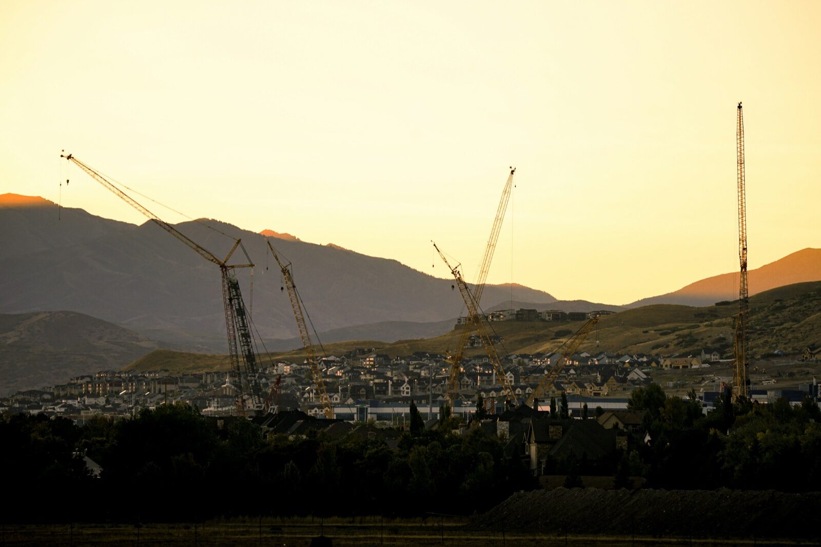 Several tall construction cranes over a building site and rows of houses at sunset, with mountains behind, in Lehi, Utah.