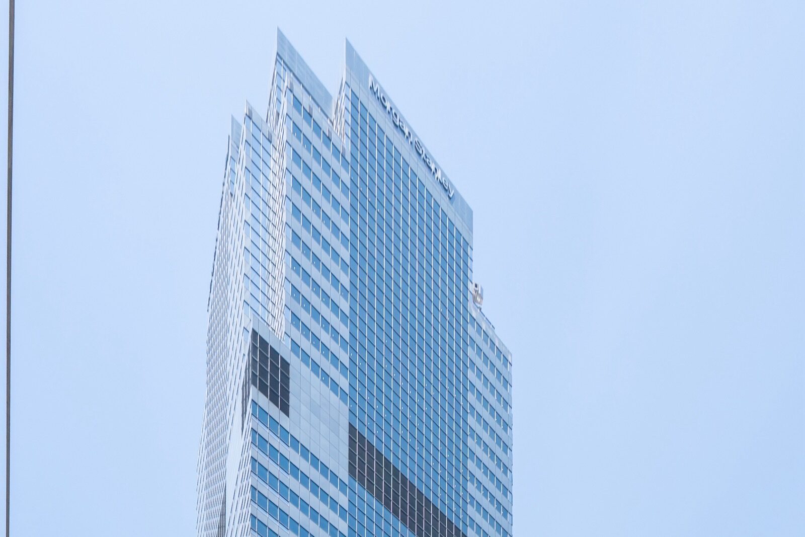 The upper floors of 1585 Broadway, Morgan Stanley's headquarters tower in Midtown Manhattan, with the Morgan Stanley name near the top of its glass facade against a pale blue sky.