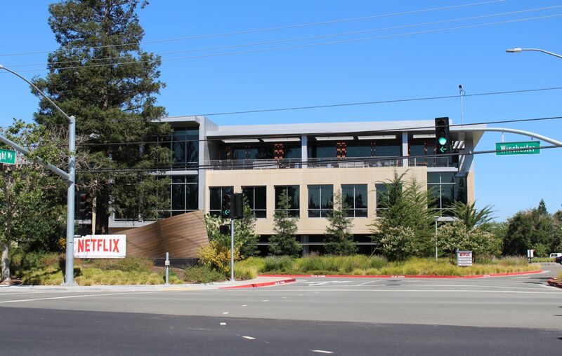 A three-storey glass and stone office building at 101 Albright Way, part of Netflix's headquarters campus in Los Gatos, California, with a red Netflix sign by the road and a traffic signal in the foreground.