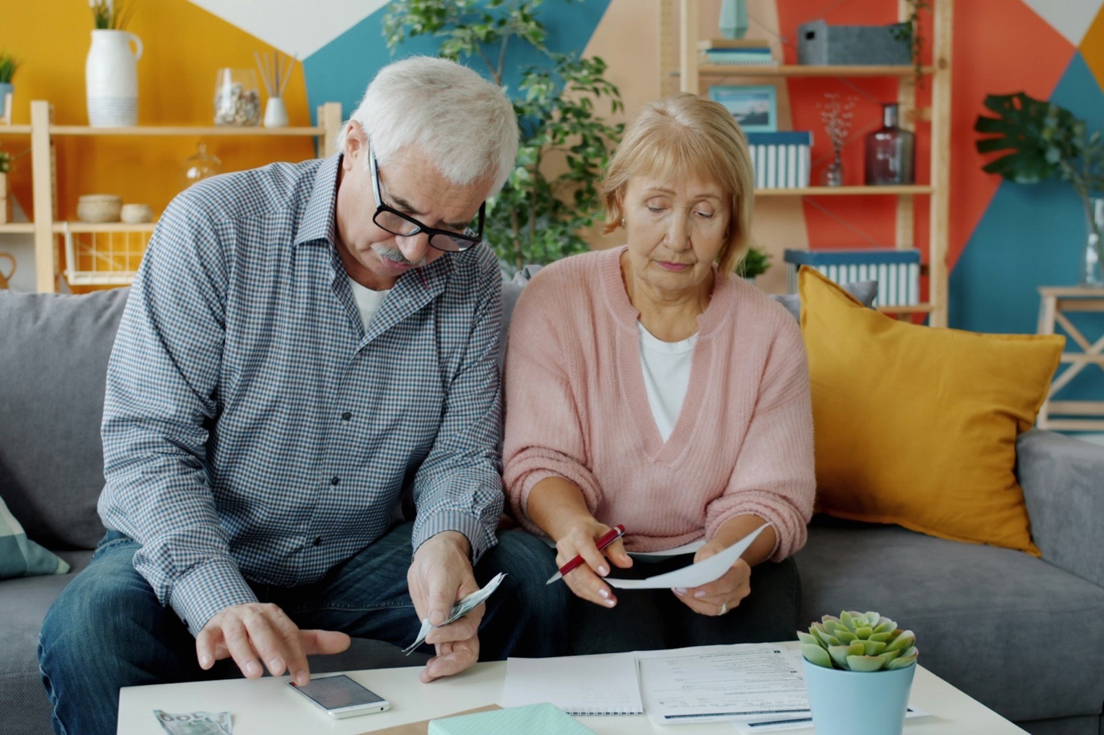 An older couple sit on a sofa going through bills and papers on a coffee table, the man holding a phone and the woman a pen.