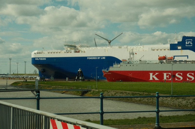Two large car-carrier ships, one blue and white and one red and white, berthed at a port behind a railing, with a wind turbine beyond.