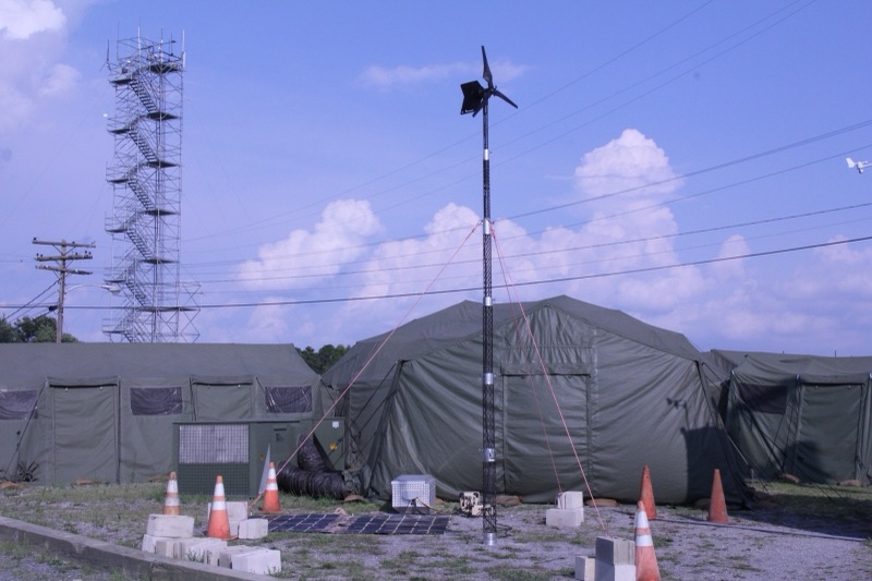 A small wind turbine on a guyed pole stands among olive-green Army tents, with a ground-level solar panel, a power unit, orange traffic cones and a steel lattice tower under a blue sky.