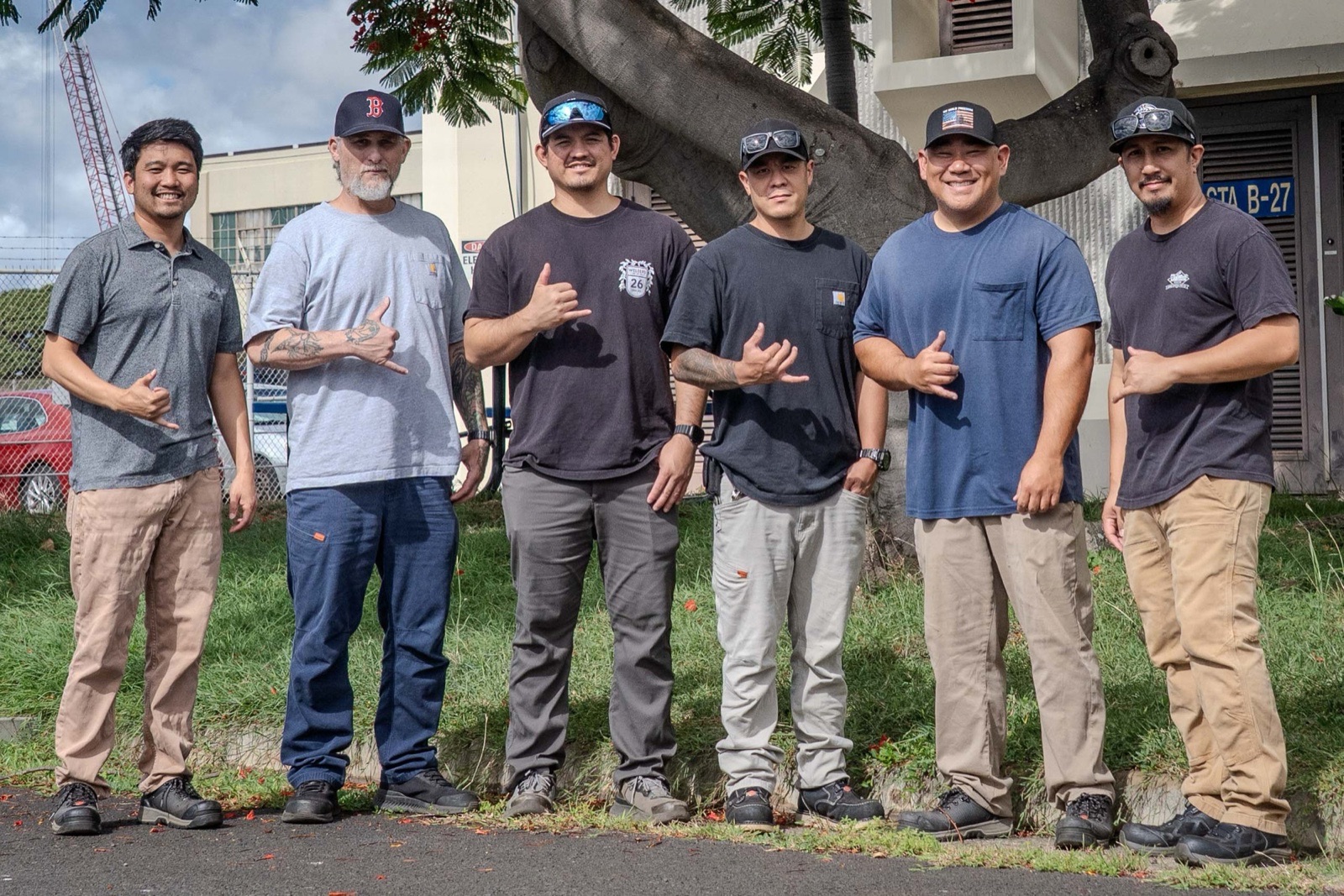 Six men in T-shirts and work trousers stand in a row on grass beneath a large tree at Pearl Harbor Naval Shipyard, smiling and making shaka hand signs.