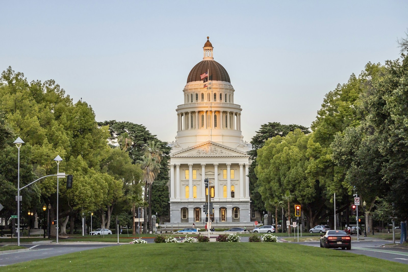 The domed California State Capitol in Sacramento at the end of a tree-lined lawn, with traffic lights in the foreground.