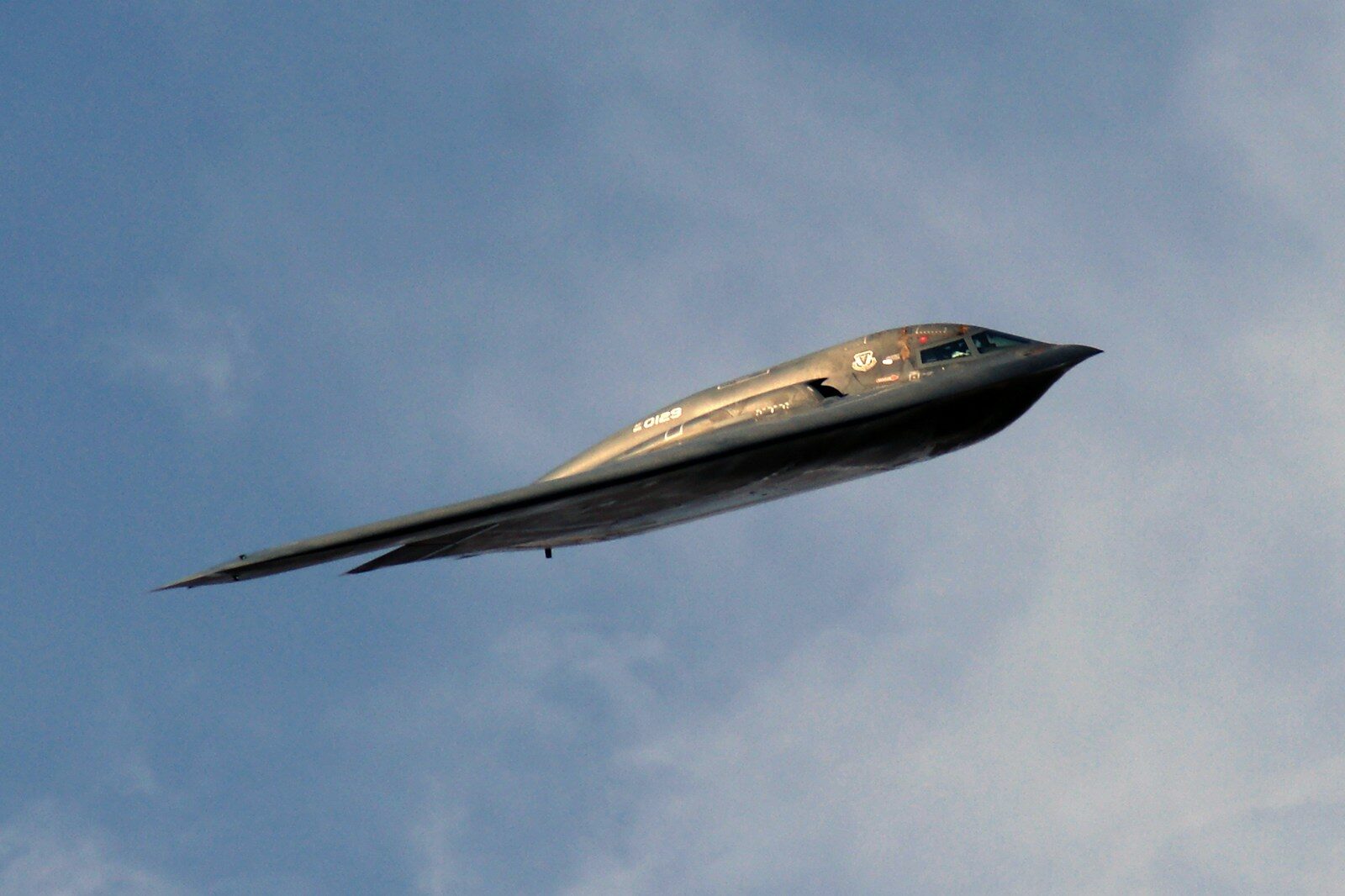 A B-2 stealth bomber in flight against a blue sky, its fuselage lit by the sun.