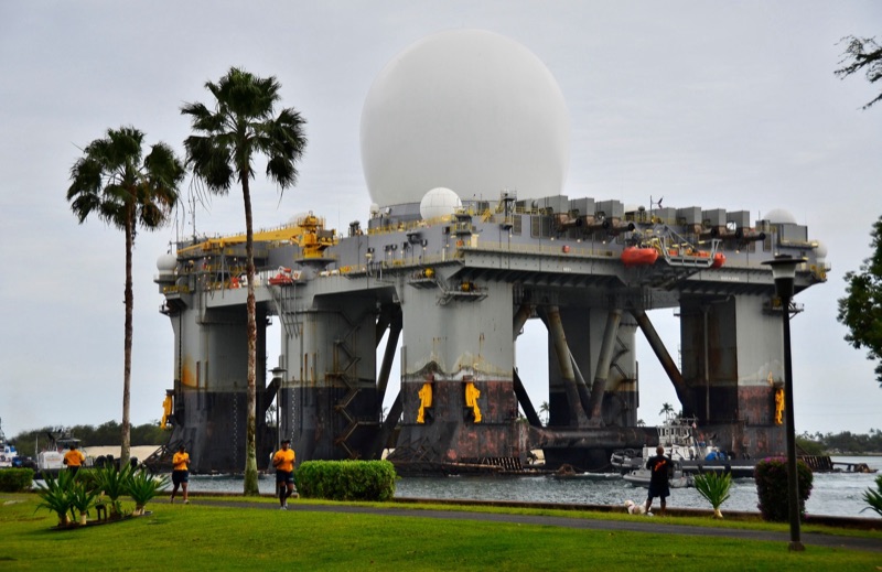 A large white radar dome on top of a semi-submersible platform, passing palm trees and lawns at Pearl Harbor.
