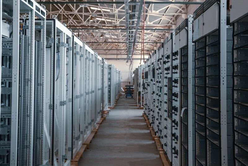 A long aisle between rows of tall grey metal equipment racks in a large industrial hall with a steel-truss roof.