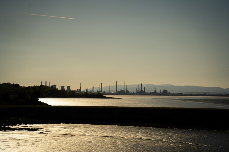 Silhouette of an oil refinery and cooling towers across an estuary at sunset, with tidal flats in the foreground.