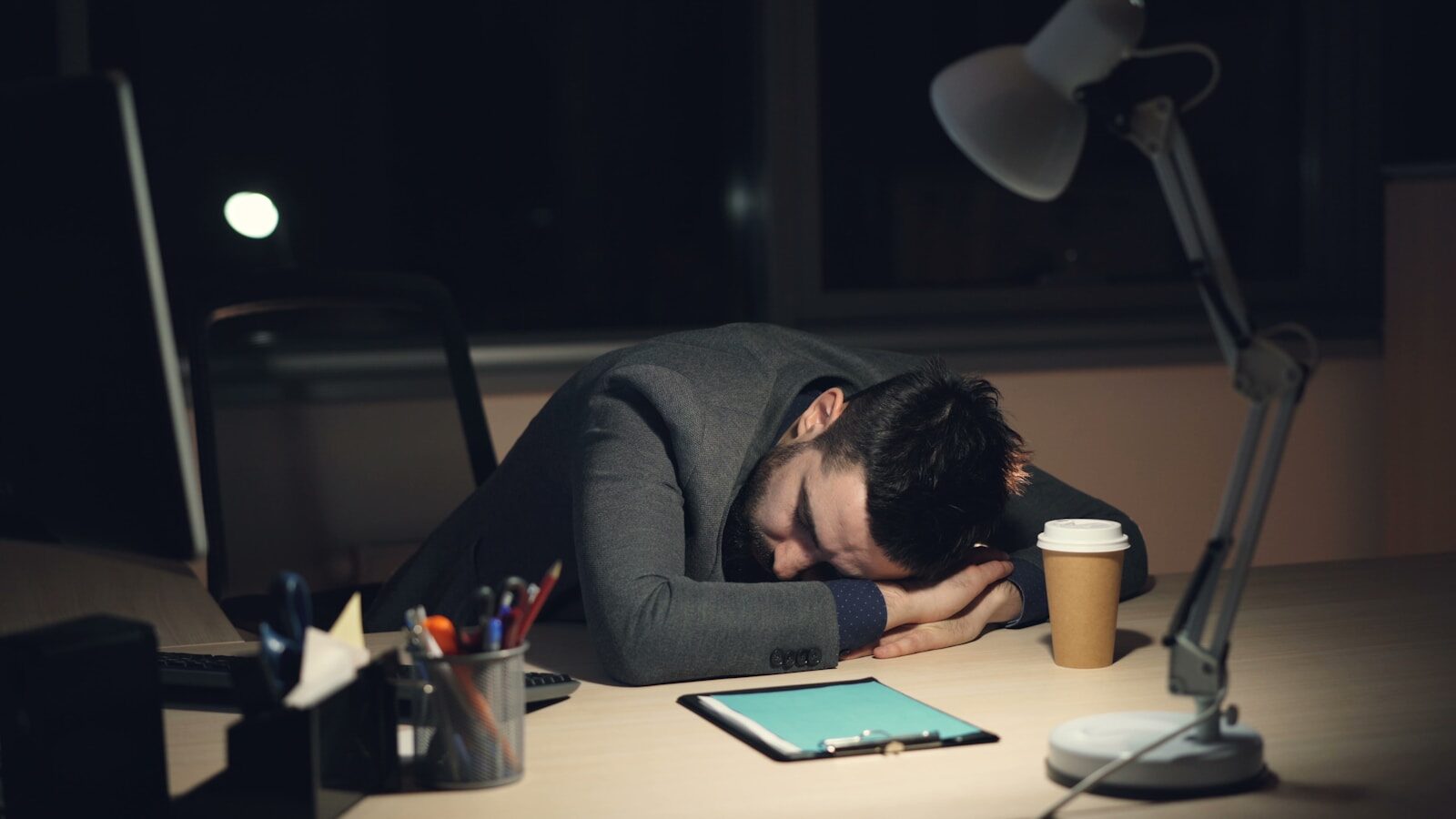 Tired guy in formal suit in sleeping on work desk in office late at night after busy working day. Turned-on computer, takeaway coffee and lamp are visible on table.