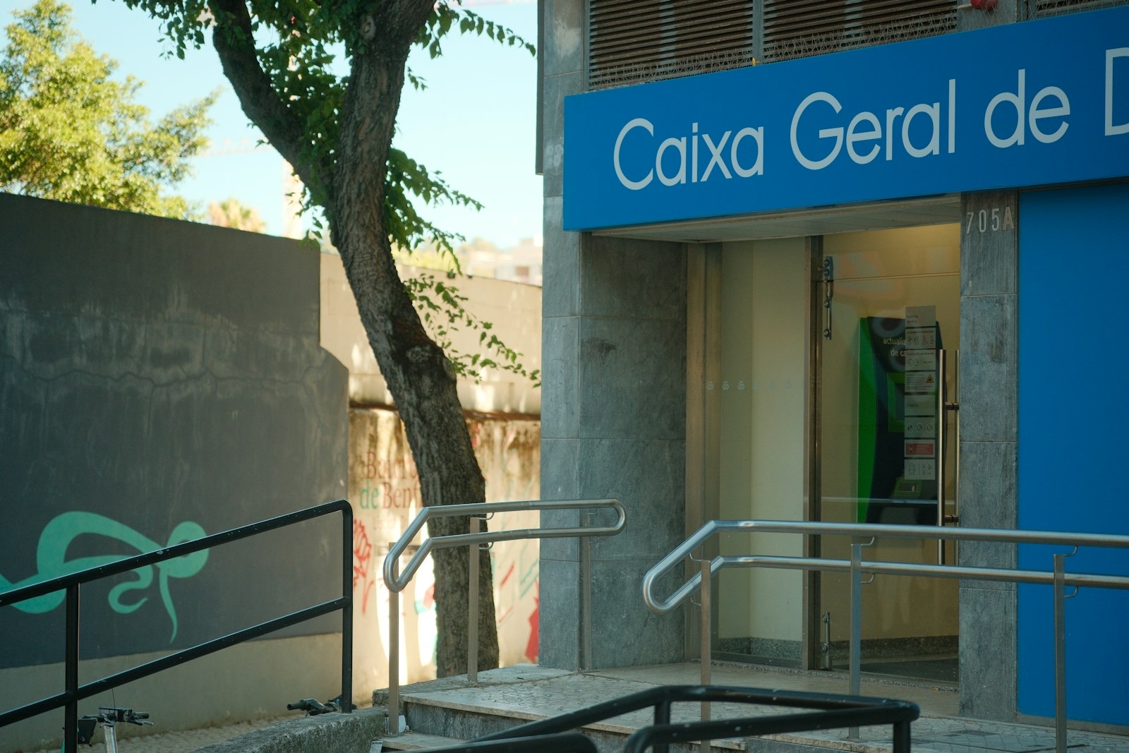 Entrance to a Caixa Geral de Depósitos bank branch in Benfica, Lisbon, framed by trees, railings and urban street art in August 2026. // Entrada de uma agência da Caixa Geral de Depósitos em Benfica, Lisboa, enquadrada por árvores, corrimões e arte urbana em agosto de 2026.