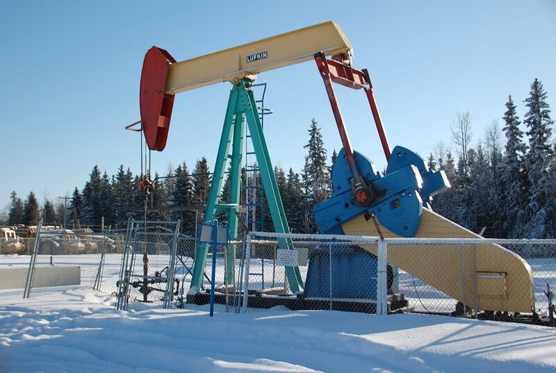 A Lufkin pumpjack with a red counterweight head, yellow beam and teal frame behind a chain-link fence on snow-covered ground, with spruce trees and a clear blue sky behind.