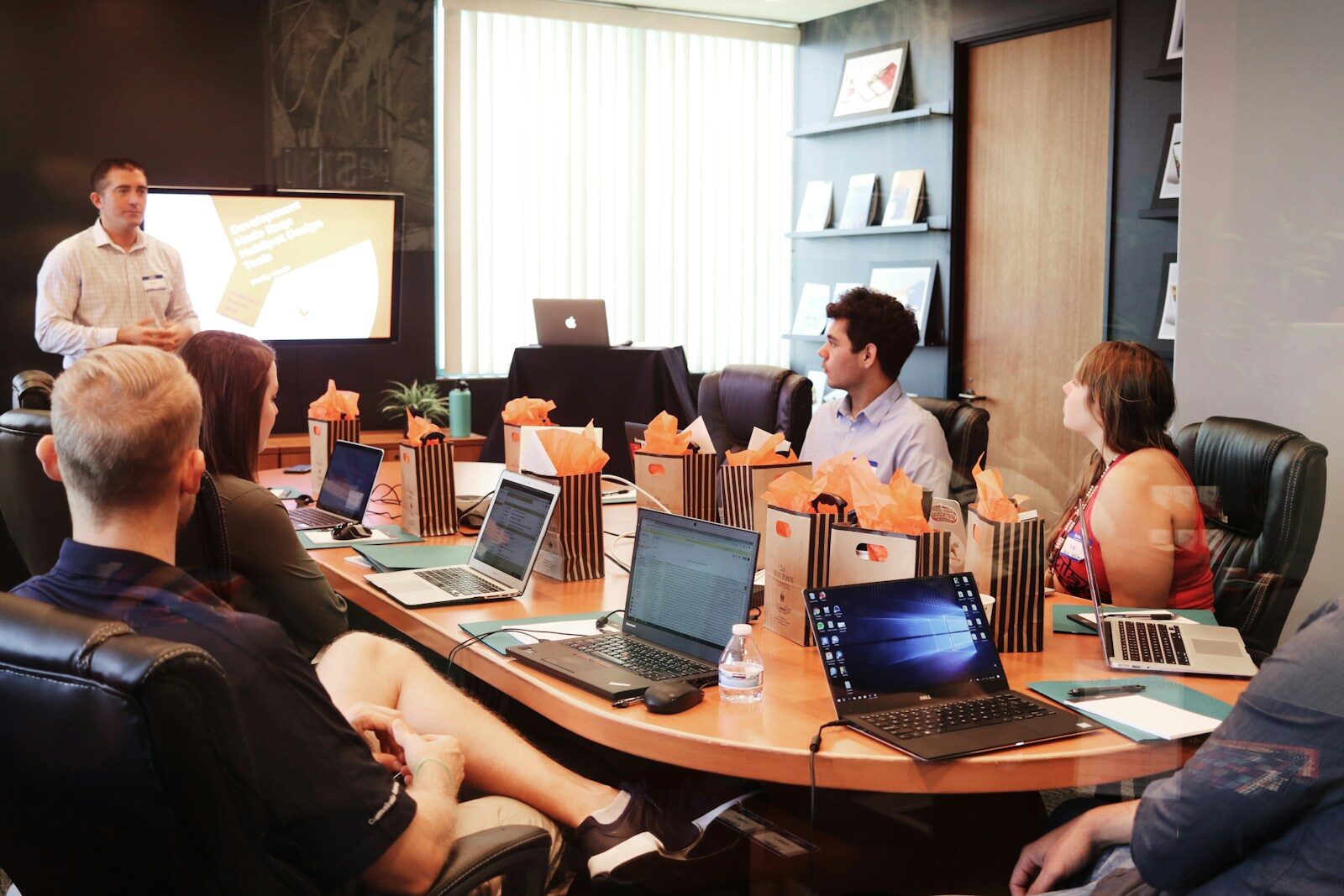 A manager standing to address colleagues seated with laptops around a table