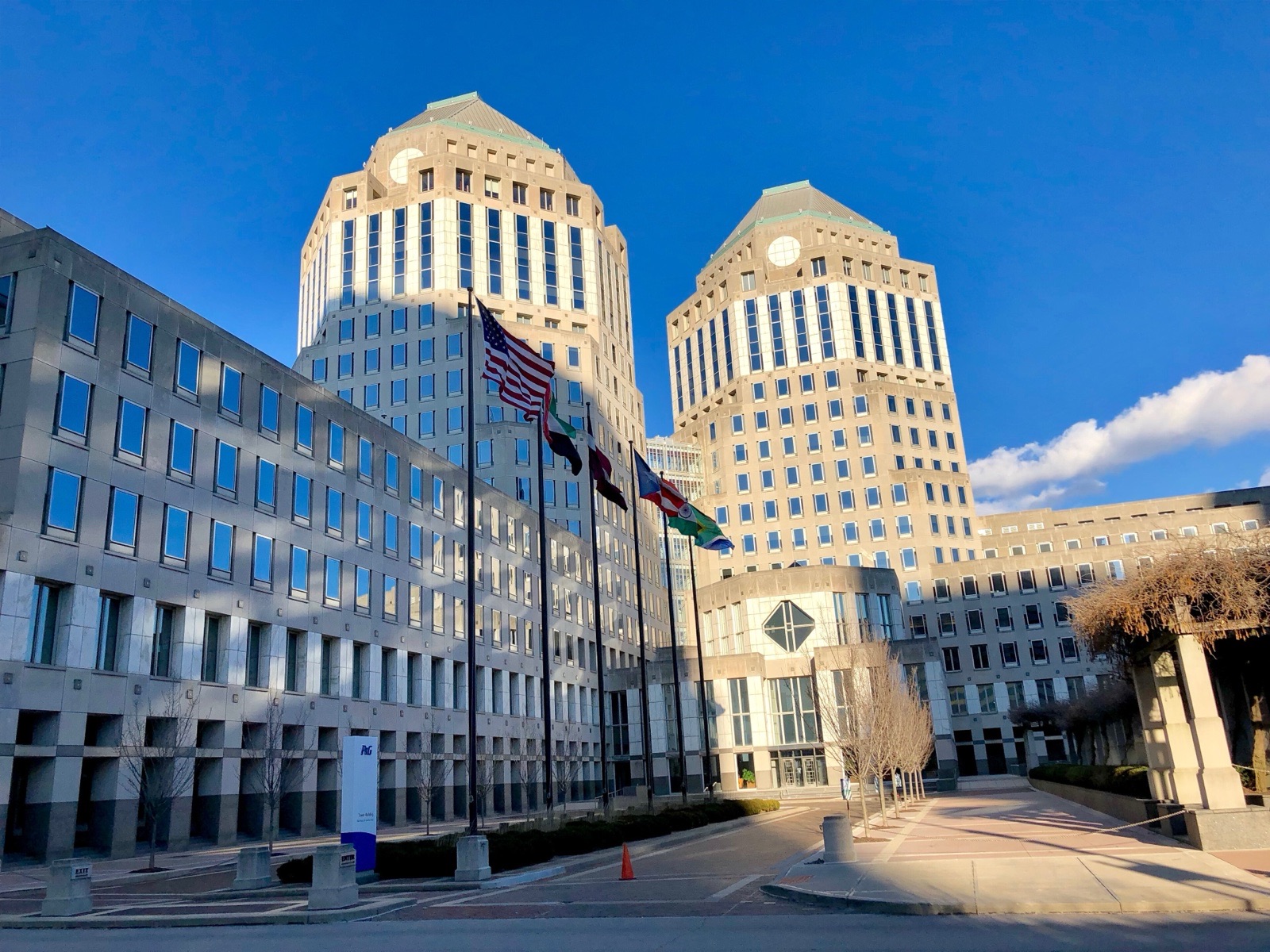 The twin limestone towers of Procter & Gamble's headquarters in Cincinnati behind a row of flagpoles and a paved plaza, under a deep blue sky.
