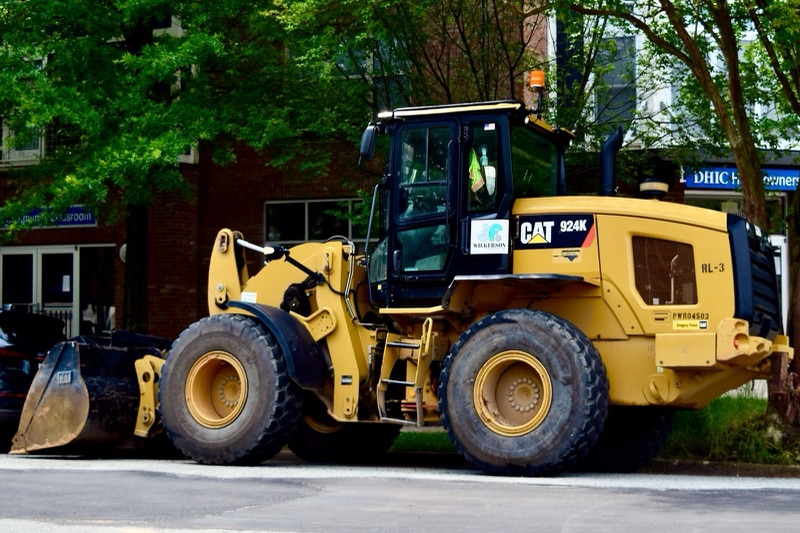 A yellow Cat 924K wheel loader parked at the kerb of a tree-lined street in front of a brick building.