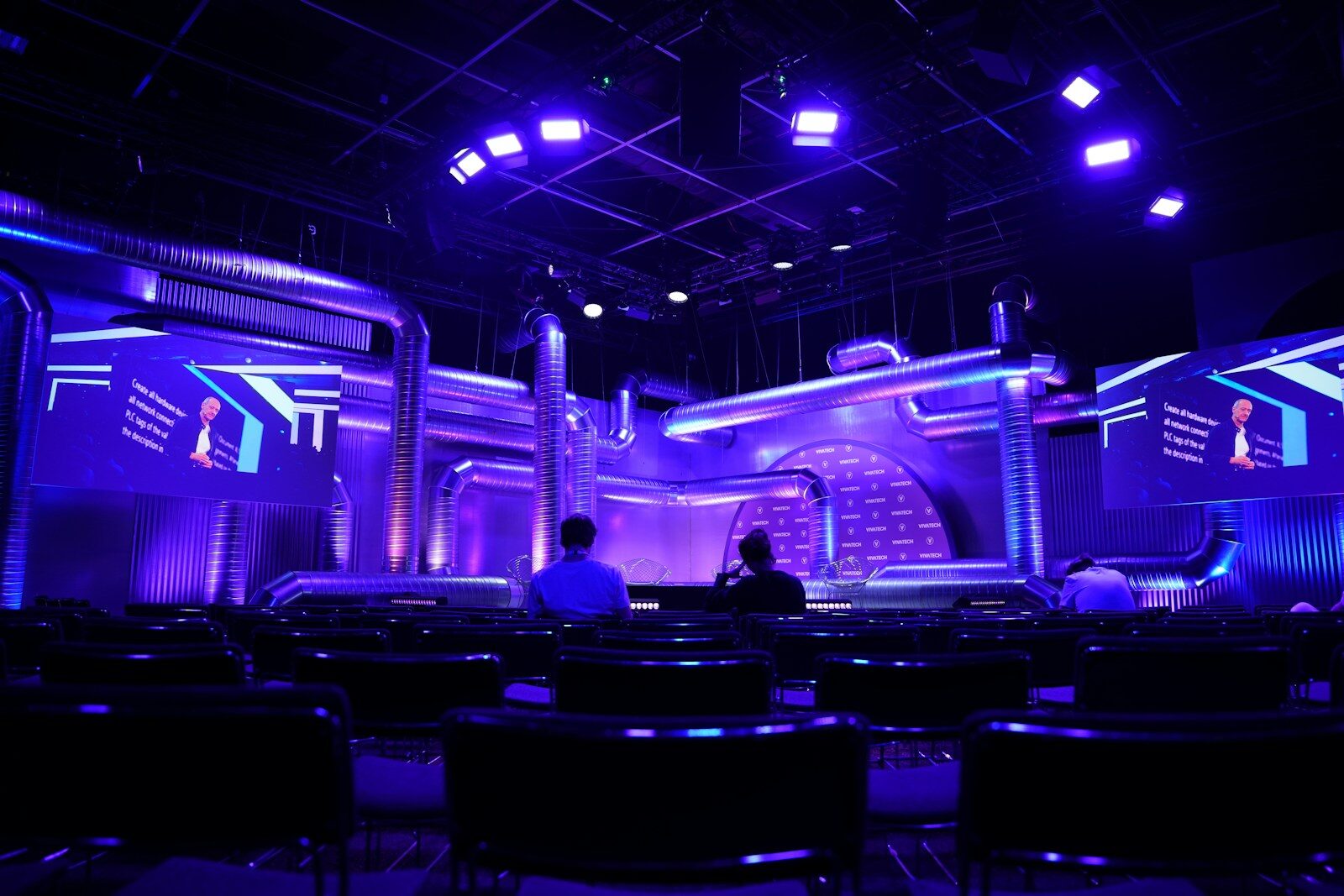 Dramatic purple and blue stage lighting illuminates a futuristic event stage set, viewed from empty auditorium seating with two silhouetted figures in the foreground. Sculptural stage design with metallic structures and screen panels creates a high-tech, sci-fi atmosphere, ideal for concepts around technology conferences, innovation events, entertainment production, and stage design.