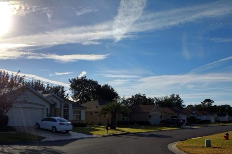A curving residential street of single-storey stucco houses with garages, palm and oak trees and trimmed lawns under a bright, streaked blue sky.