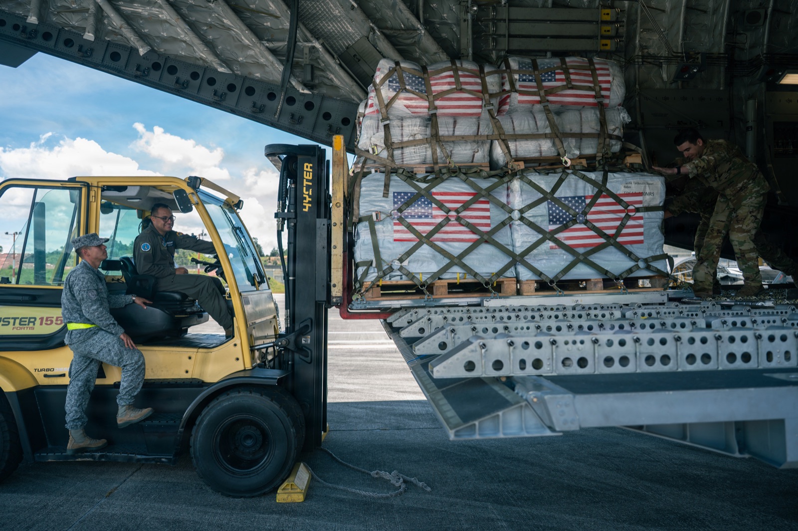 Flag-marked pallets of relief supplies coming off the ramp of a military transport aircraft onto a forklift.