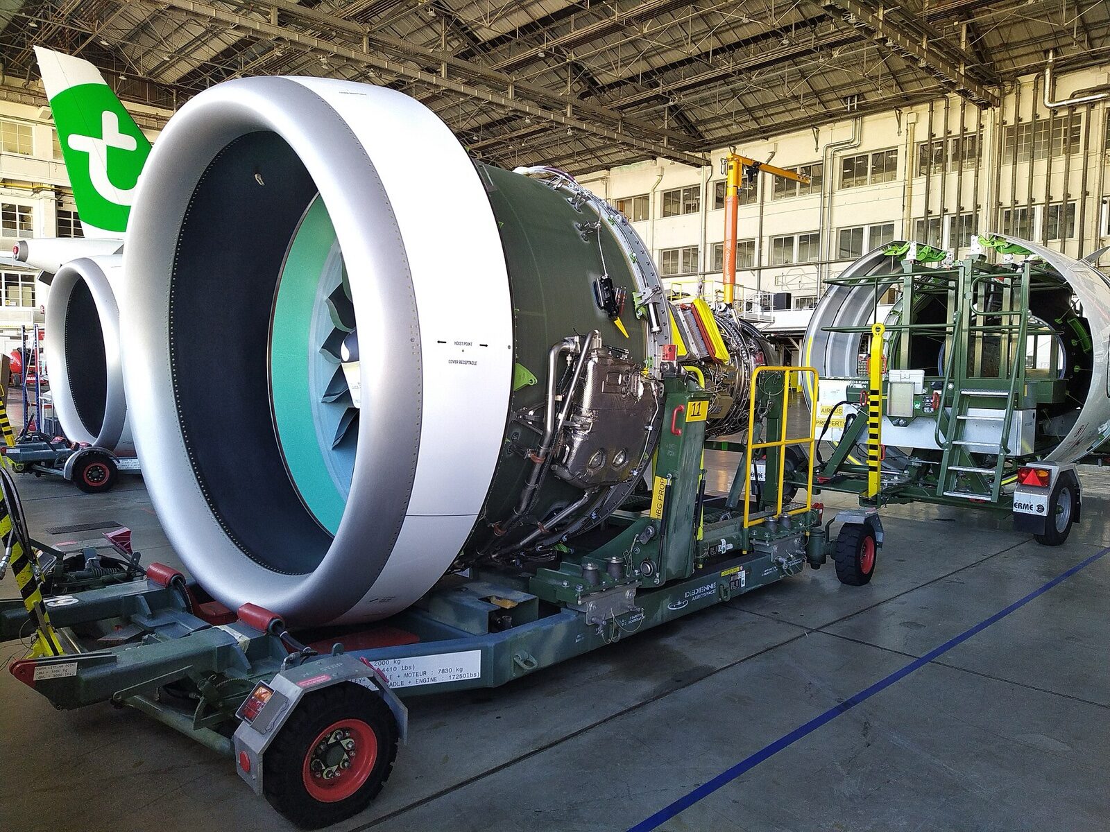 A Pratt & Whitney PW1000G geared turbofan engine, its cowling open, on a transport stand inside an aircraft hangar, with a second engine and an airliner tail behind it.