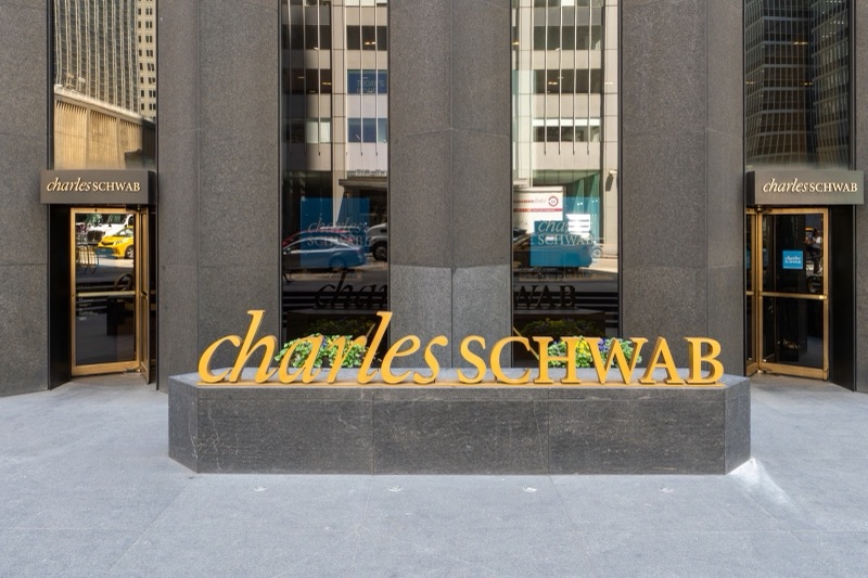 Gold Charles Schwab lettering on a granite planter outside the firm's office at the base of the CBS Building on Sixth Avenue in New York, with glass doors and dark granite piers behind.