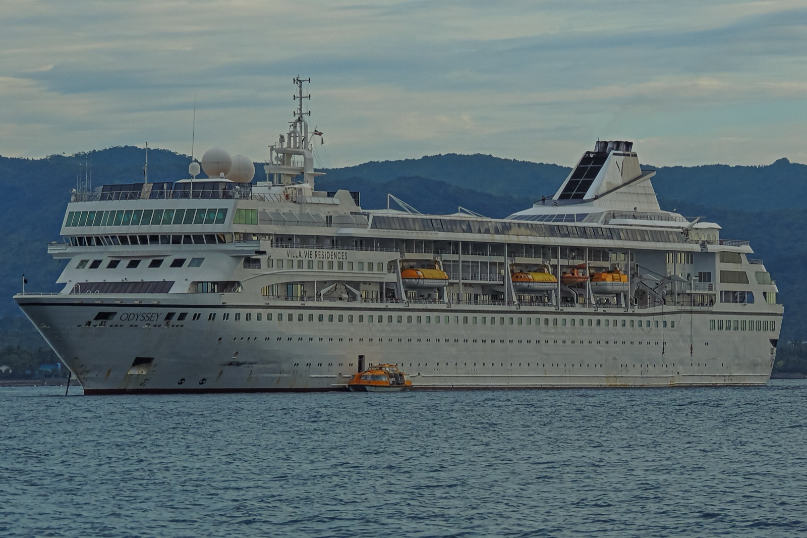 A white cruise ship with Odyssey and Villa Vie Residences painted on its side lies at anchor beside a small orange tender, with green hills behind.