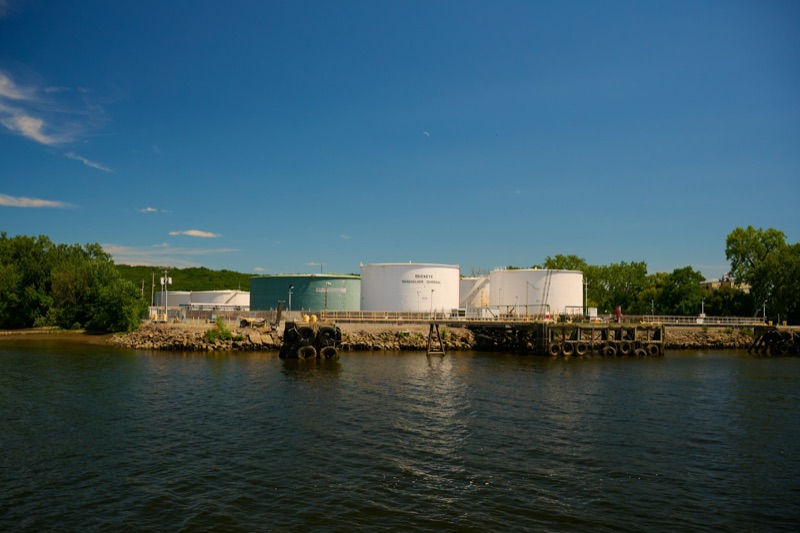 White fuel storage tanks at a riverside terminal under a blue sky, seen from the water.