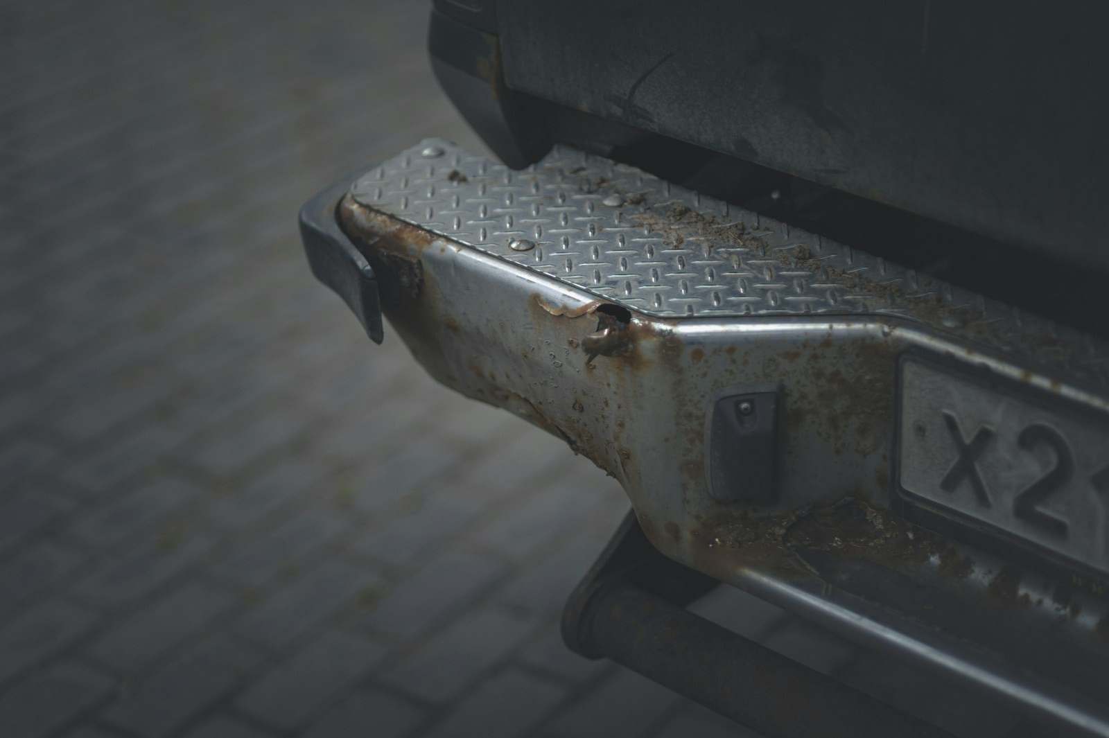 The rusted, dented rear bumper of an old vehicle parked on a cobbled street.