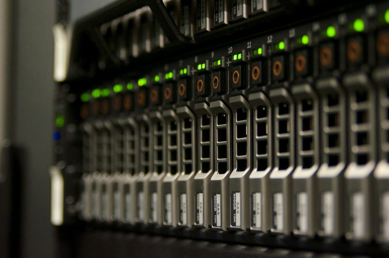 The front of a rack-mounted server photographed at an angle, showing a row of grey hard-drive bays with green status lights glowing above them in a darkened equipment room.