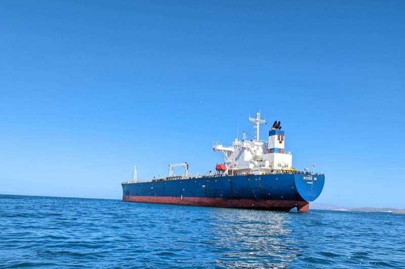 A crude oil tanker with a dark blue hull and red waterline lies at anchor on calm blue water under a clear sky, its white superstructure at the stern. No other vessels are visible.