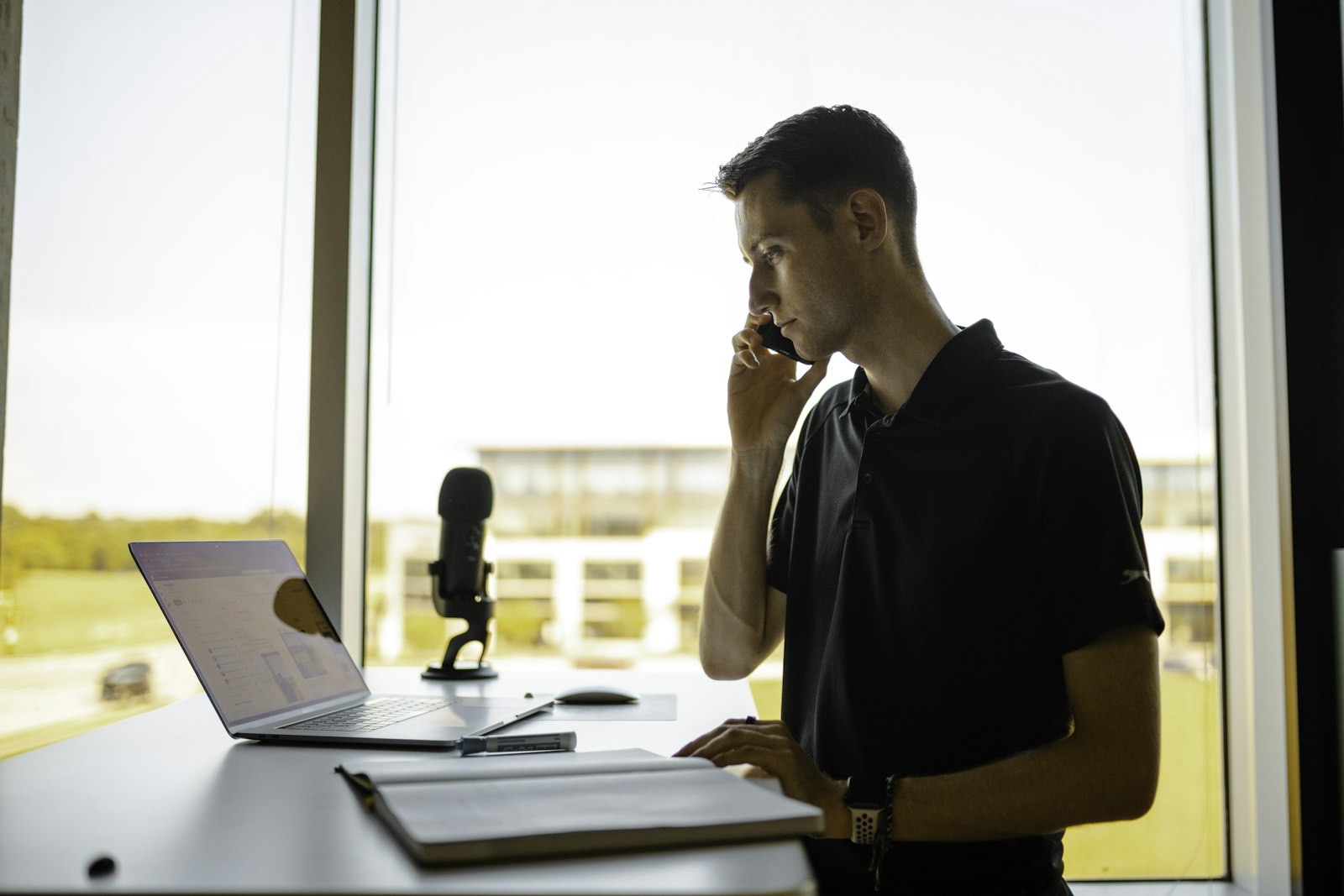 A man at a desk working on a computer while using a phone.