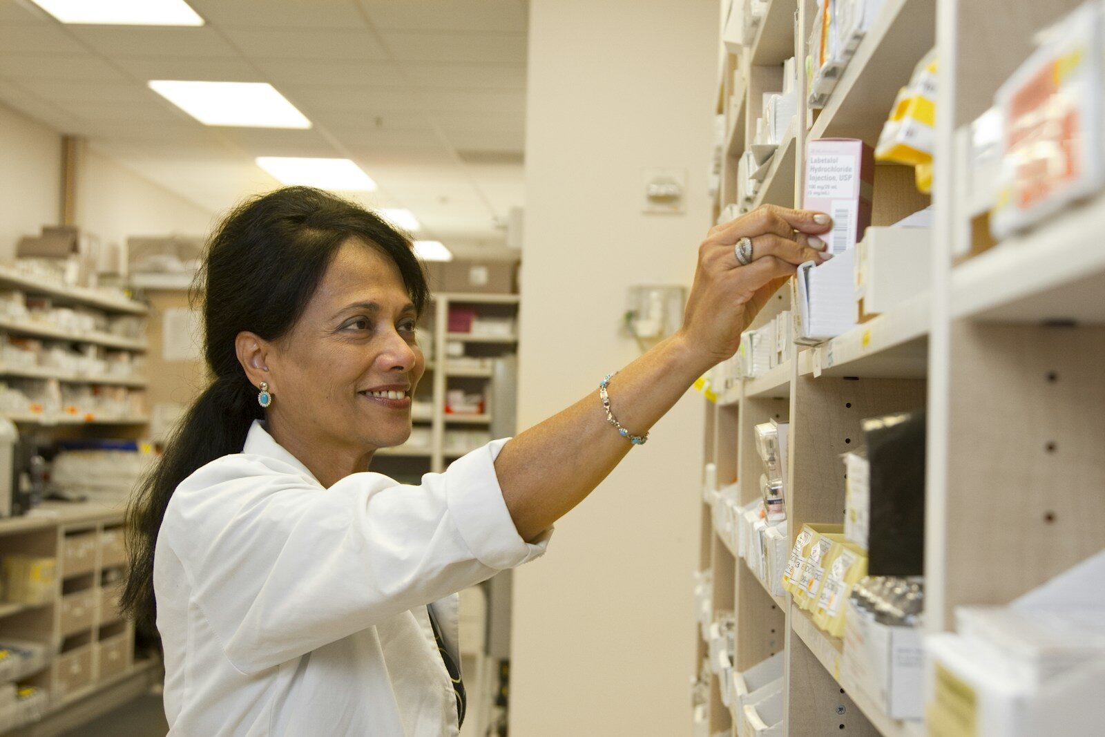 A pharmacist reaching for a box of injectable medication on a hospital pharmacy shelf.