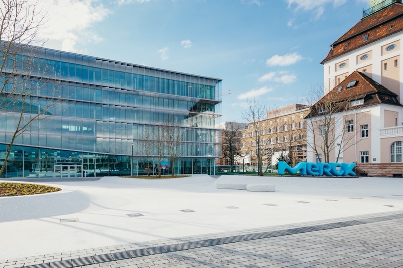 A glass office building and an older building with a tiled roof around a paved plaza, with a large blue Merck sign standing on the plaza.