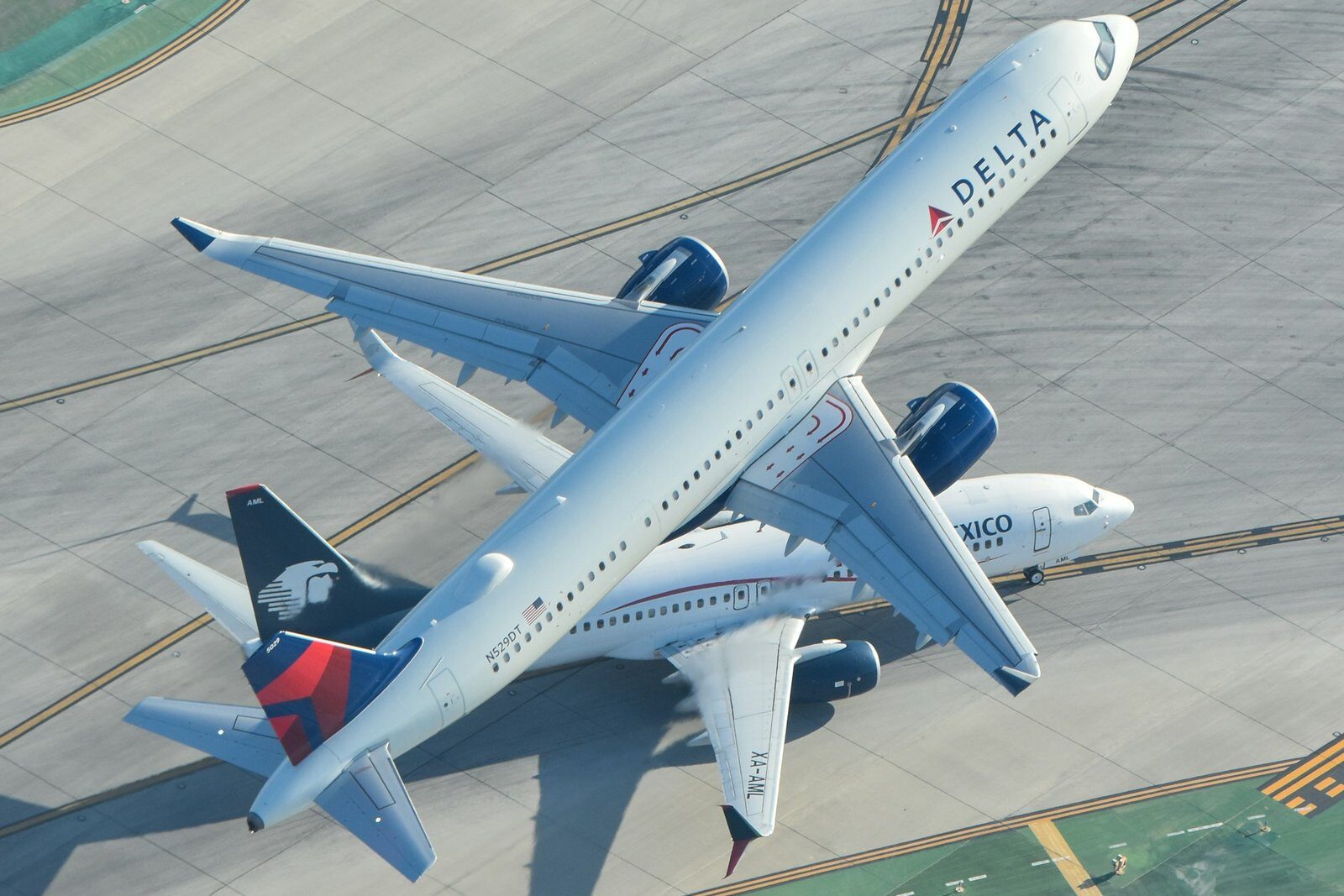 Seen from above, a white Delta Air Lines jet with a red and blue tail flies low over an airport taxiway, passing over an Aeromexico airliner on the ground.
