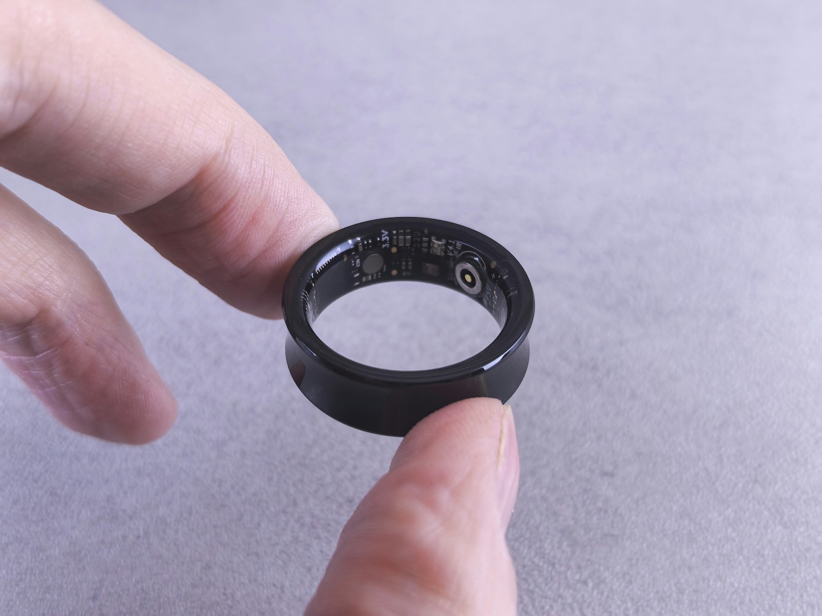 A black smart ring held upright between a thumb and forefinger against a grey surface, with the sensor array and circuit board visible on the inner surface of the band.