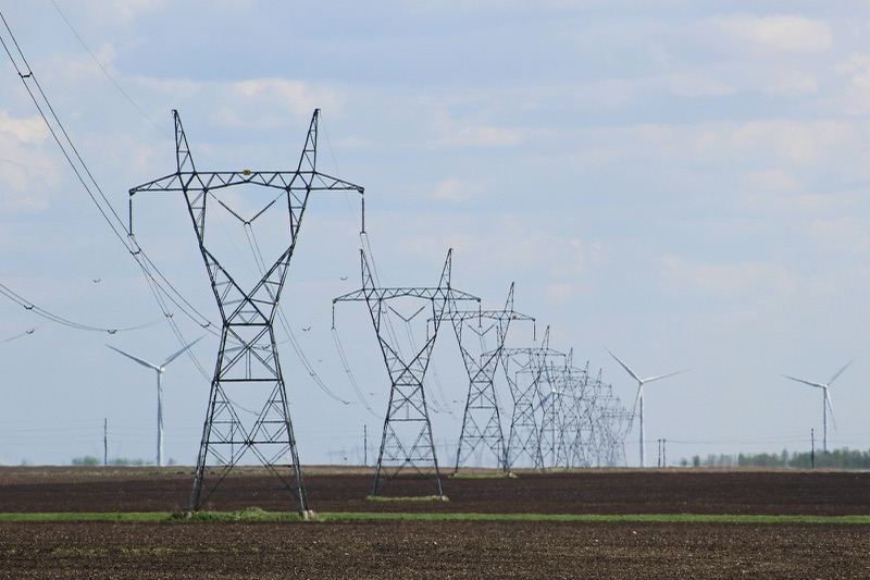 A line of steel lattice transmission towers carrying high-voltage lines across flat, freshly tilled farmland, with wind turbines on the horizon.