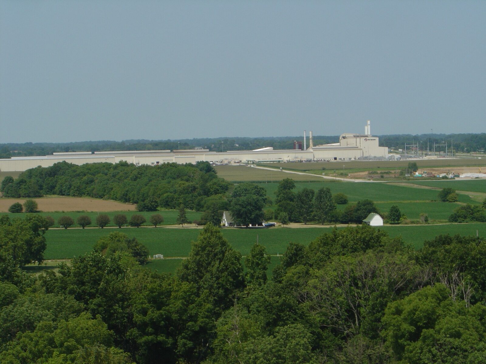 The long, low white buildings and stacks of a steel mill on the horizon beyond farm fields and trees.