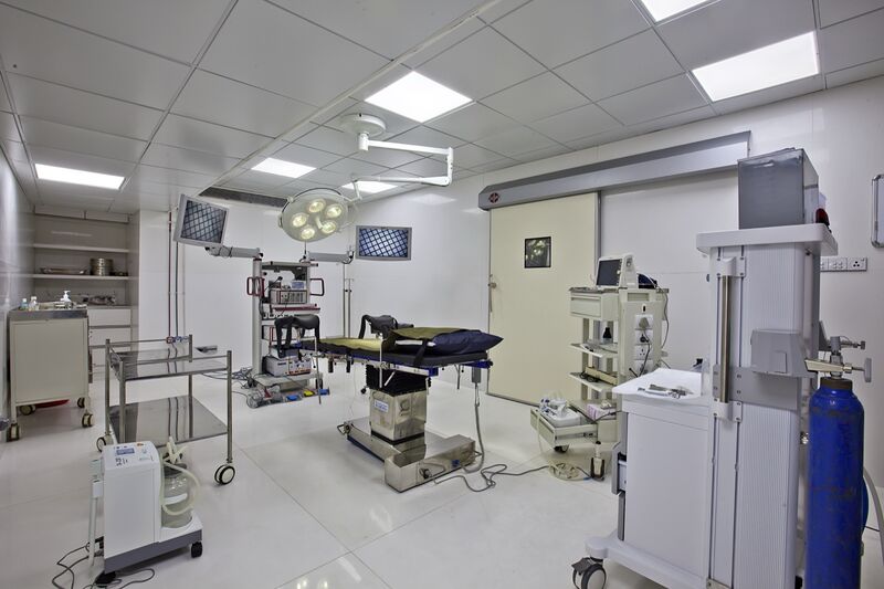 An empty operating theatre with a padded surgical table under a ceiling-mounted surgical light, flanked by two wall monitors, equipment carts, an anaesthesia unit and a blue gas cylinder, under white ceiling panels.