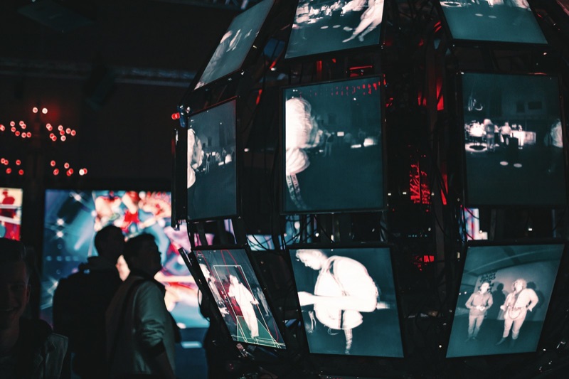A cluster of video screens mounted on a multi-sided tower in a darkened hall, each panel showing a different image, with the silhouettes of several people watching from the left.